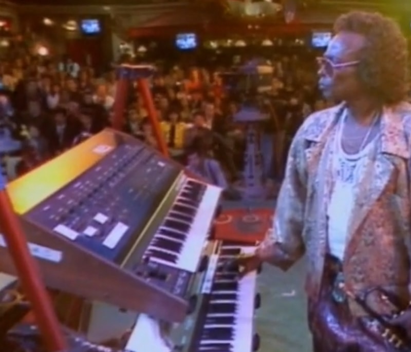 Photo of Miles Davis on stage in front of a keyboard rack with the audience in the background.